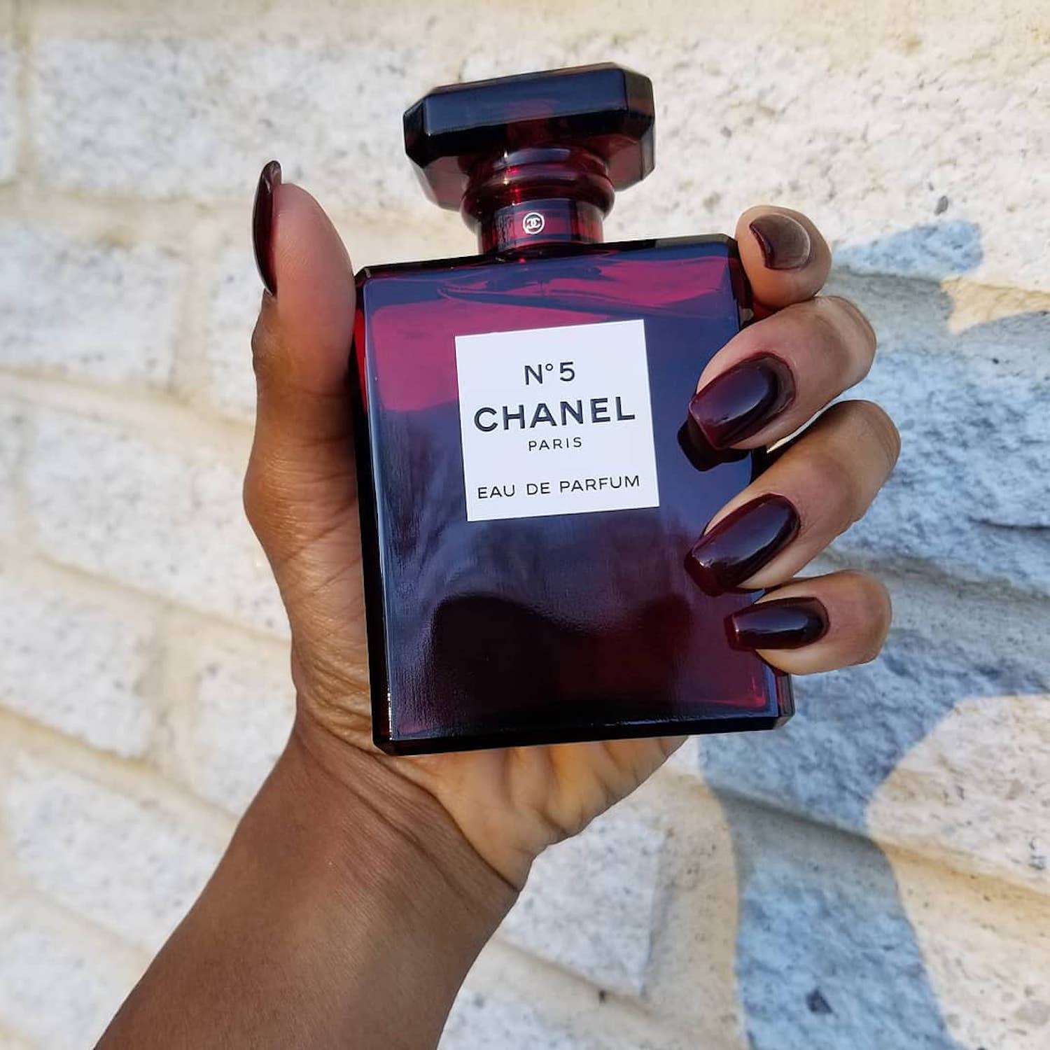 Uma mão com pregos de caixão pretos da cor vermelho sangue segura uma garrafa vermelha de Chanel No. 5
