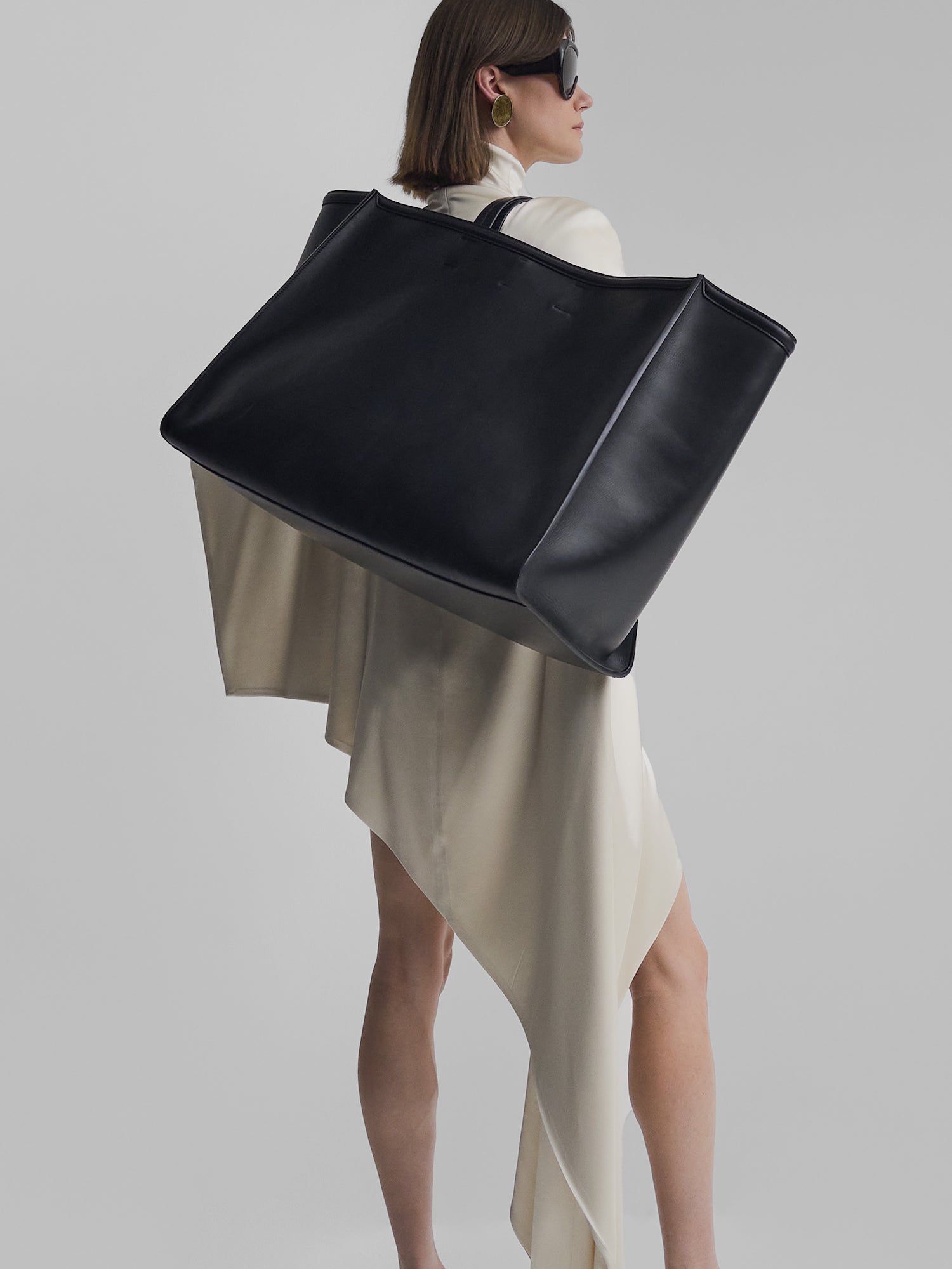 Modelo com uma bolsa preta Phoebe Philo XL Caba em um vestido branco assimétrico e óculos de sol grandes