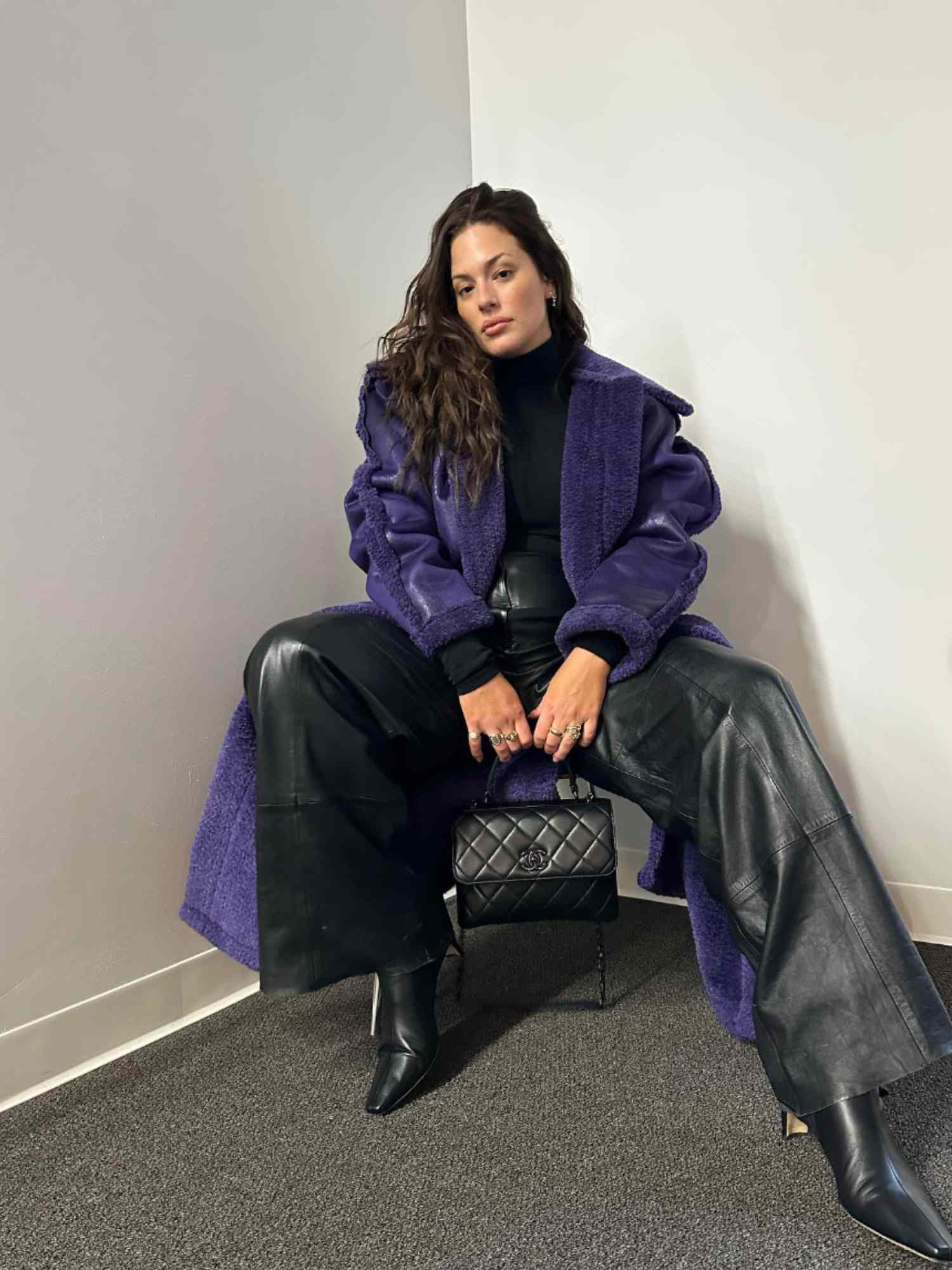Ashley Graham segura uma bolsa Chanel preta acolchoada enquanto veste um casaco de pele roxo, calça preta com detalhes em couro, gola alta preta e botas pretas