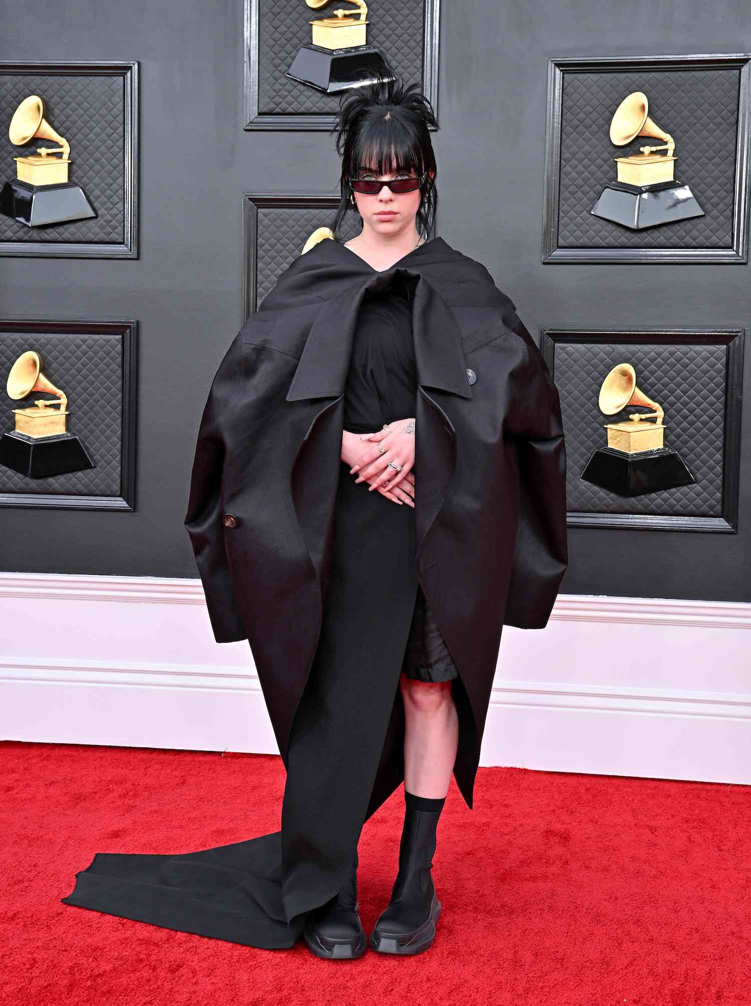 Billie Eilish usa vestido preto com capa Rick Owens, botins plataforma e óculos escuros no Grammy Awards de 2022