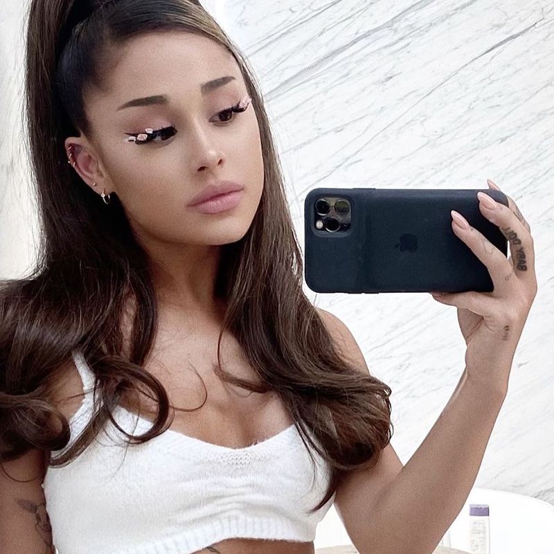 Ariana Grande posa para selfie espelhado com tatuagem no dedo e cílios florais
